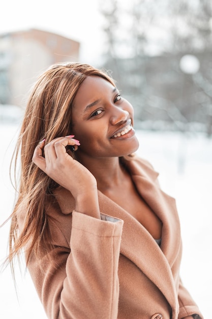 Stylish beautiful young happy african woman with a smile in a\
beige coat walks on the street with snow