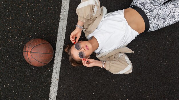 Stylish beautiful young hipster girl in fashionable sportswear with a windbreaker Tshirt and leggings is wearing sunglasses and lies resting on the pavement