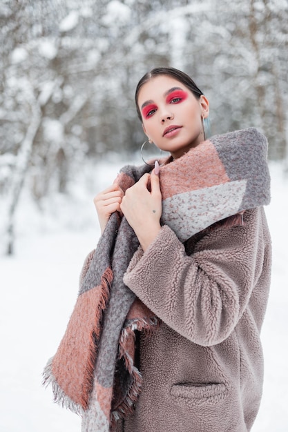 Stylish beautiful young Scandinavian woman in fashionable clothes with a warm coat and scarf in a winter forest in Norway