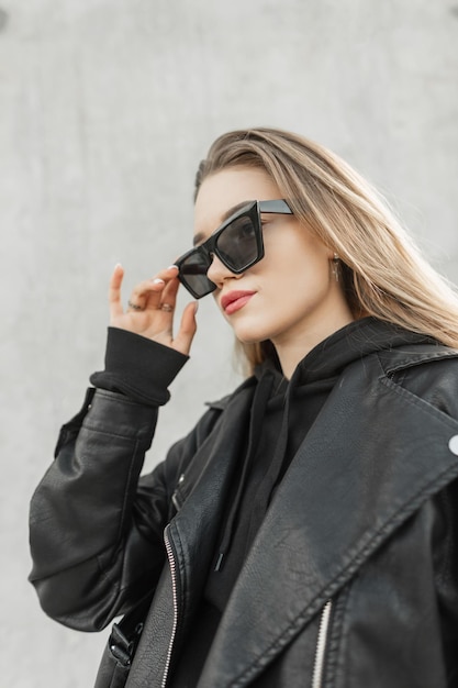 Stylish beautiful young woman in fashionable casual clothes with leather jacket and hoodie wears sunglasses and walks on the street near the gray wall Fashion style and beauty
