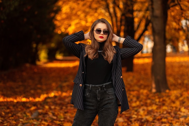 ジャケット、セーター、サングラスとファッショナブルな秋の服を着たスタイリッシュな美しい若い女性は、明るい秋の色の葉のある公園でポーズをとります。女性のビジネススタイルと美しさ