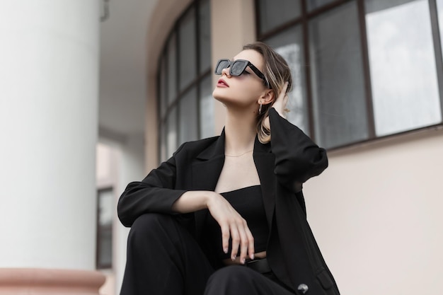 Stylish beautiful young woman model with black sunglasses in fashion black business outwear with a jacket sits in the city Female trendy elegant clothes look