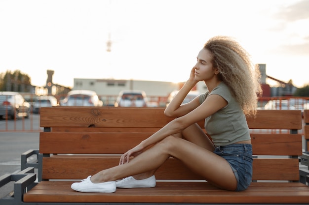 白いスニーカーとファッショナブルなデニムのショートパンツで巻き毛のスタイリッシュな美しい若い女性モデルは、日没時にベンチに座っています