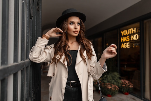 Stylish beautiful young woman with curly brunet hair in a fashionable hat with a stylish beige coat walks on the street near a cafe. Elegant feminine fall style and beauty