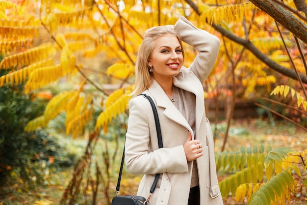 黄色い紅葉の近くの流行のコートに笑顔でスタイリッシュな美しい若い女性