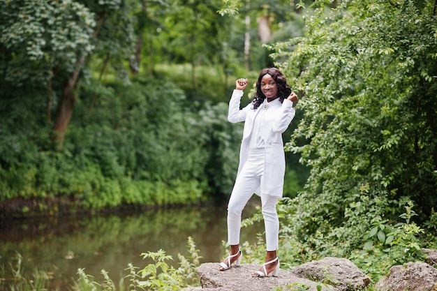 Ragazza afroamericana nera alla moda che posa alla pietra sul fiume del fondo del parco