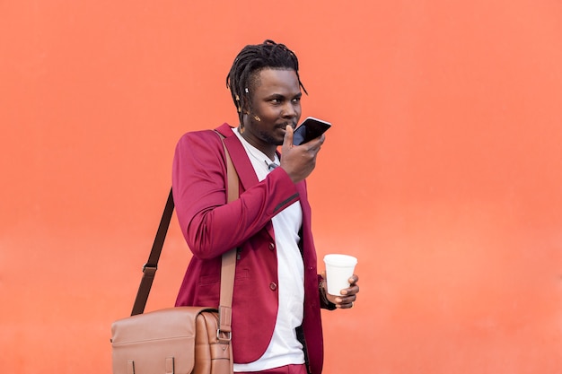 ブリーフケースとコーヒーを身に着けたスタイリッシュな黒人実業家は、赤い背景、テキスト用のコピースペース、テクノロジーとコミュニケーションの概念の前で彼のスマートフォンに音声メッセージを録音します