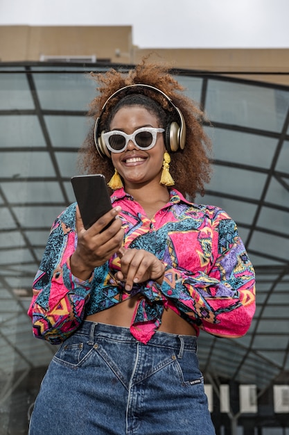 スマートフォンの屋外でインターネットをサーフィンヘッドフォンでスタイリッシュな黒人女性