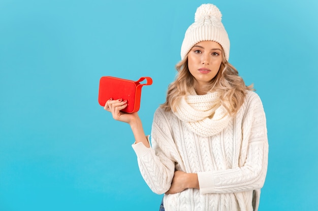 Стильная белокурая красивая молодая женщина, держащая беспроводной динамик, слушает музыку в белом свитере и вязаной шапке, позирует моды в зимнем стиле, изолирована на синей стене