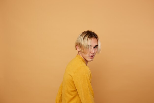 Stylish blond guy dressed in yellow sweater is posing on the beige background in the studio .