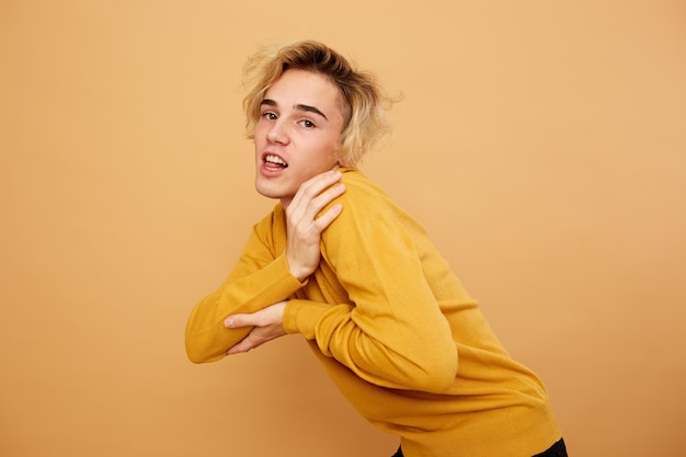 Stylish blond guy dressed in yellow sweater is posing with his hand on his head on the beige background in the studio .