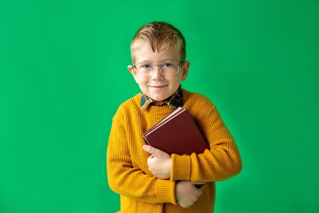 彼の手で開いた本を持つスタイリッシュな金髪の学齢期の少年黄色のセーターと緑の背景の分離のメガネで陽気な少年知識の力を学ぶ学校教育に戻る