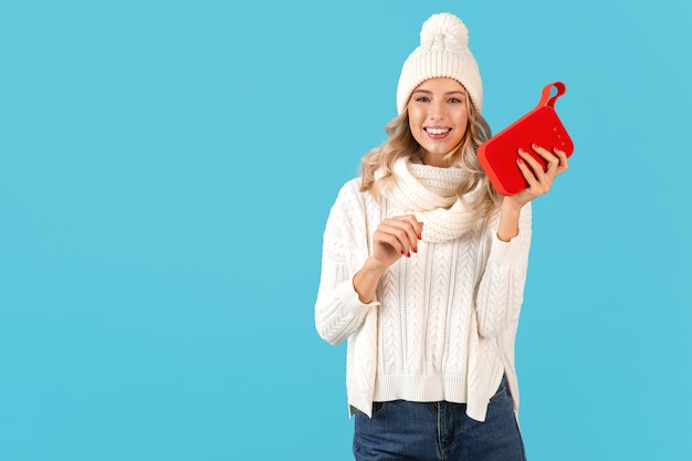 Stylish blond smiling beautiful young woman holding wireless speaker listening to music happy dancing wearing white sweater and knitted hat winter style fashion posing isolated on blue wall