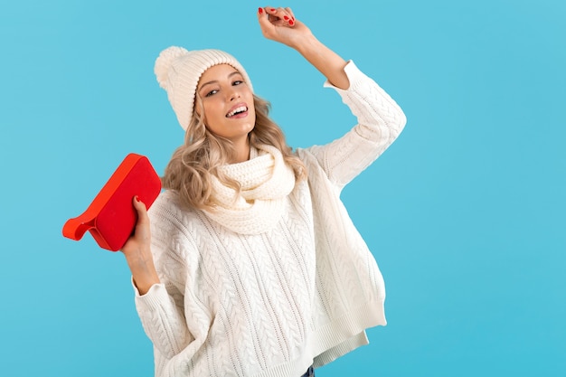 Stylish blond smiling beautiful young woman holding wireless speaker listening to music happy dancing wearing white sweater and knitted hat winter style fashion posing 