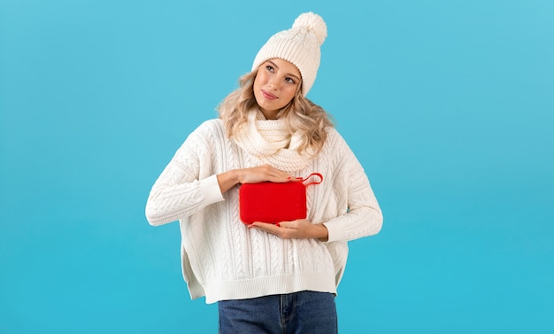 Bella giovane donna sorridente bionda alla moda che tiene altoparlante senza fili che ascolta la musica maglione bianco da portare felice e cappello lavorato a maglia che posa sull'azzurro