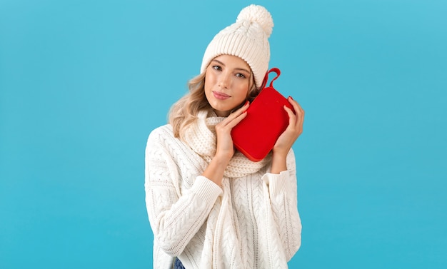 Stylish blond smiling beautiful young woman holding wireless speaker listening to music happy wearing white sweater and knitted hat winter style fashion posing isolated on blue background