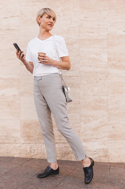 stylish blond woman wearing white t-shirt using cell phone, while standing against beige wall outdoor in summer and looking aside at copyspace with takeaway coffee