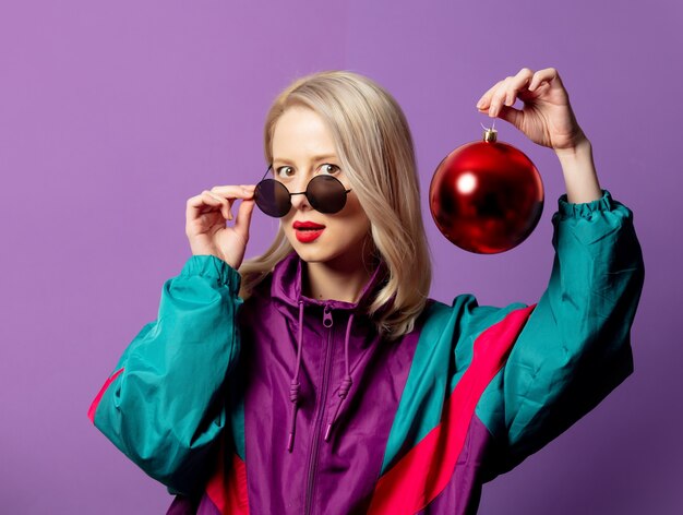 Stylish blonde in 80s windbreaker and roud sunglasses holds Christmas bauble on purple wall