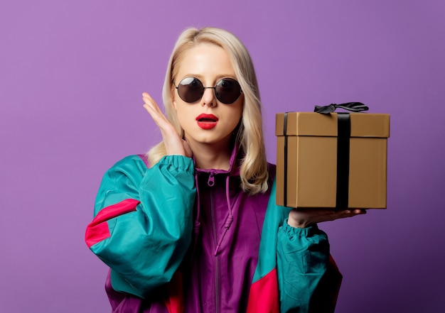 Stylish blonde in 80s windbreaker and round sunglasses with gift box on purple wall