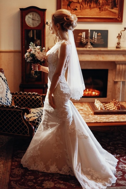 Photo stylish blonde bride walking with bouquet to groom in the morning in hotel wedding day concept beautiful woman back in white dress with flowers in vintage room