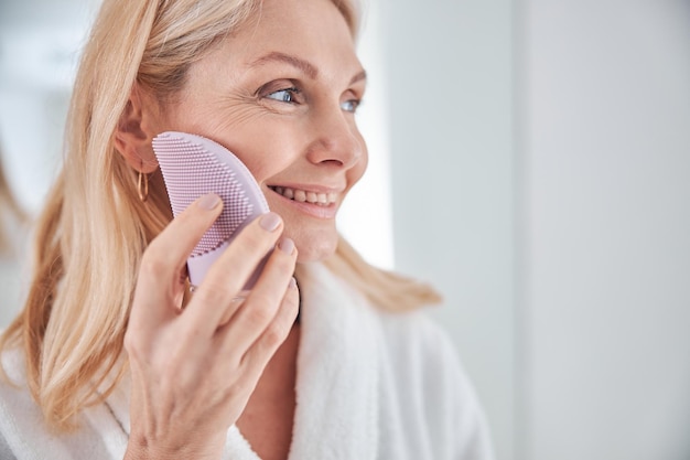 Stylish blonde Caucasian female with eclectic facial massager in hand in bathroom