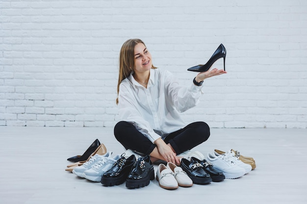 Photo stylish blonde choosing shoes while sitting on the floor side view of a lot of womens shoes on the floor a choice of stylish shoes