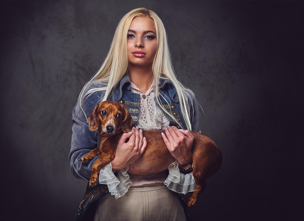 A stylish blonde female dressed in old fashioned jacket holds a red badger dog.