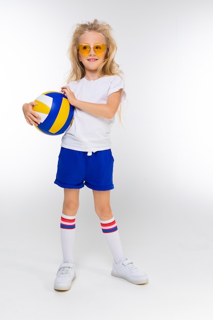 Stylish blonde girl in sports shorts, a T-shirt and gym shoes with glasses stands with a basket ball on a white wall