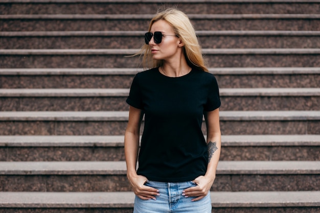 Stylish blonde girl wearing black t-shirt and glasses posing against street