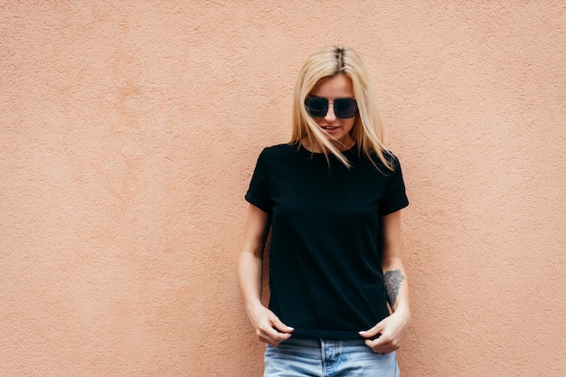 stylish blonde girl wearing black t-shirt and glasses posing against wall