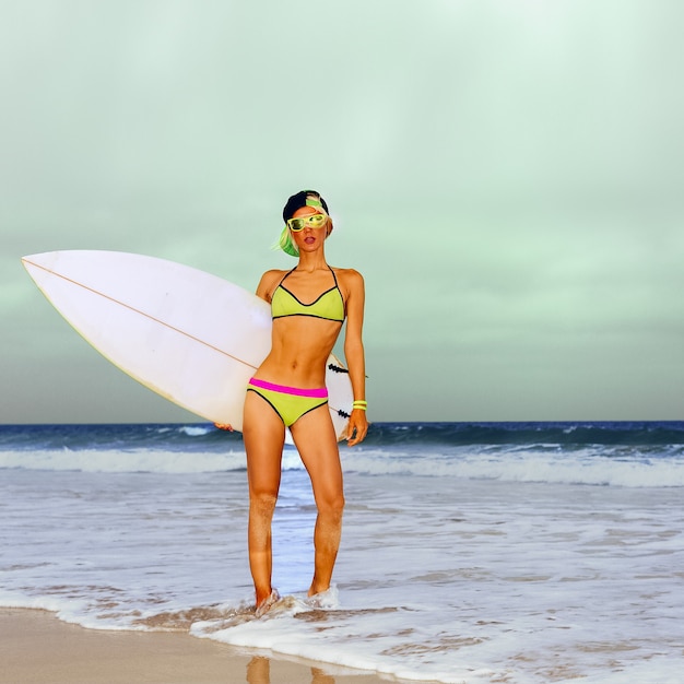 Стильная блондинка стоя на пляже с доской для серфинга.