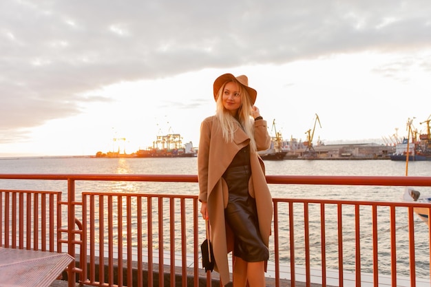 Stylish blonde woman in felt hat and coat at sunrise at sea