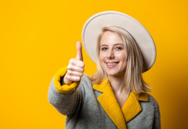 Stylish blonde woman in hat and seasonal coat giving thumb up