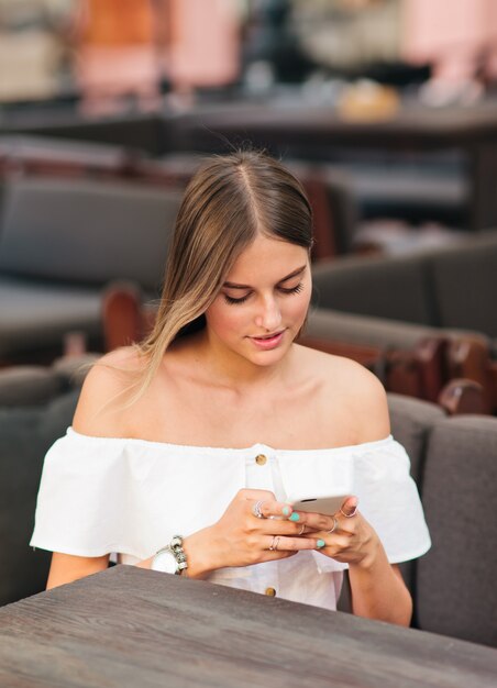 Стильная блондинка держит смартфон в руках, сидя за столиком в уличном кафе