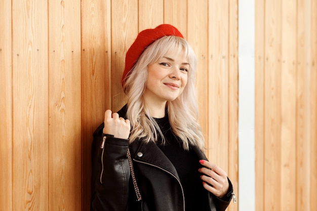 Stylish blonde woman in red beret and leather jacket in front of wooden wall