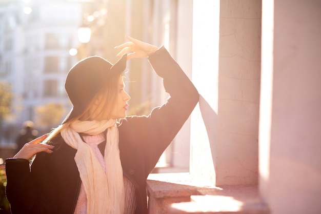 Stylish blonde woman wearing coat and hat posing at the old street. Space for text