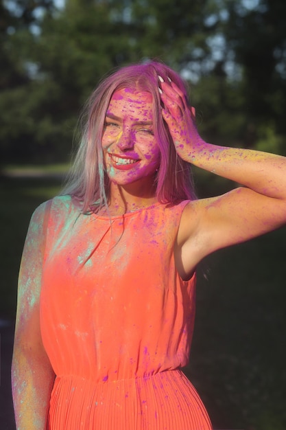 Stylish blonde woman wearing orange dress posing covered with a colorful dry paint at the park