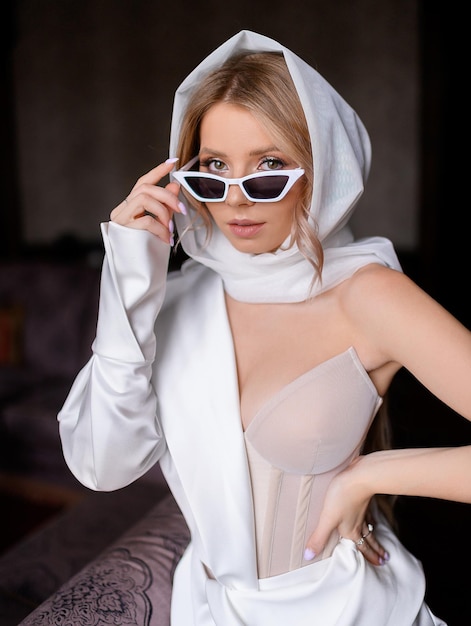 Stylish blonde young woman posing in sunglasses and white silk scarf on hand Fashion girl caucasian model dressed in beige corset standing indoor Retro styling Beauty Sixties style Retro woman