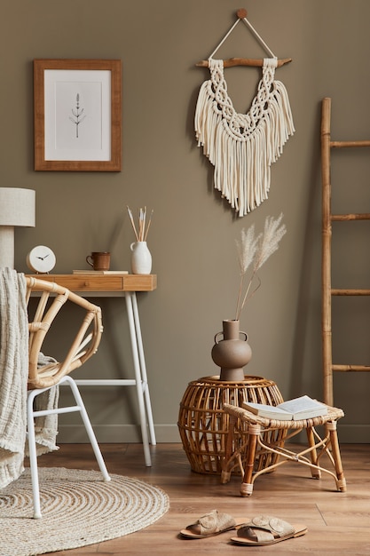 Stylish bohemian interior of home office space with wooden desk, rattan armchair, brown frame, macrame, office supplies, lamp, decoration and elegant personal accessories in home decor.