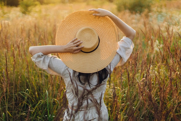 Stylish boho woman with straw hat posing among wild grasses in sunset light back view Summer