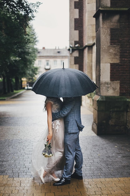 Stylish bride and groom kissing under umbrella on background of old church in rain Provence wedding