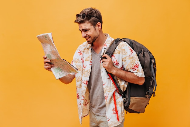 Stylish bright man with sunglasses in white printed clothes holding large backpack and looking at map on orange wall