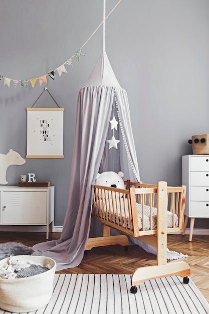 Arredamento scandinavo elegante e luminoso della stanza del neonato con poster, mobili di design bianchi, giocattoli naturali, baldacchino grigio sospeso con culla in legno, leggio, accessori e orsacchiotti