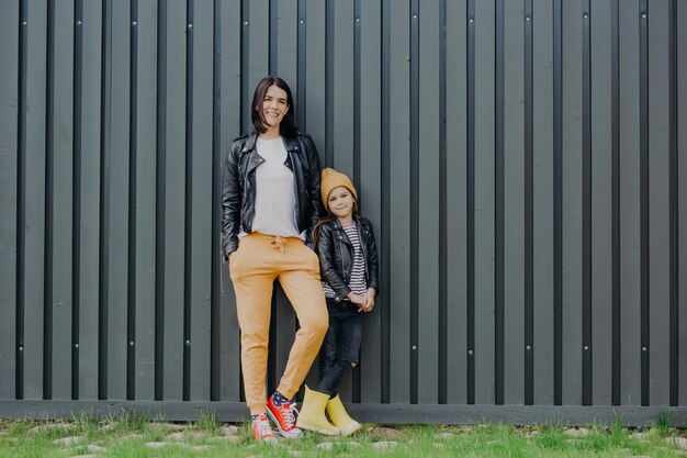 Stylish brunette female dressed in fashionable leather jacket and sport trousers and her little daughter stands near pose at camera against fence background have walk outdoor during weekend