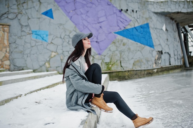 Stylish brunette girl in gray cap casual street style on winter day against colored wall