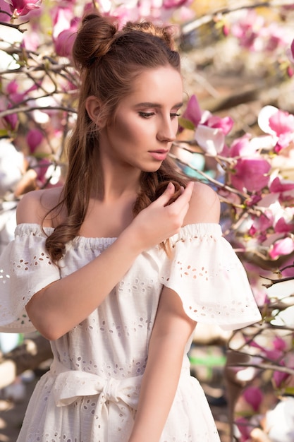 Stylish brunette girl posing near the flowering magnolia tree in white lace dress