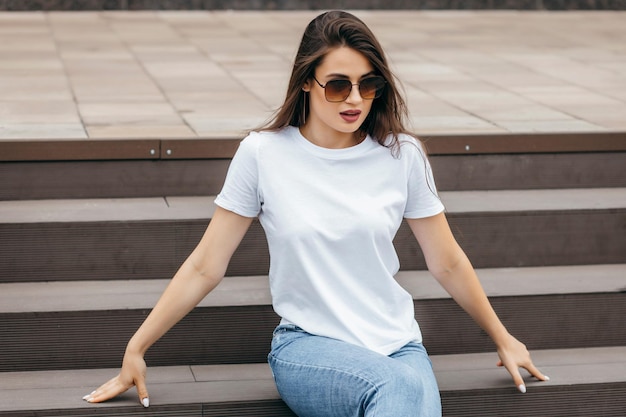 Stylish brunette girl sitting on the steps wearing white tshirt blue jeans and glasses posing agai