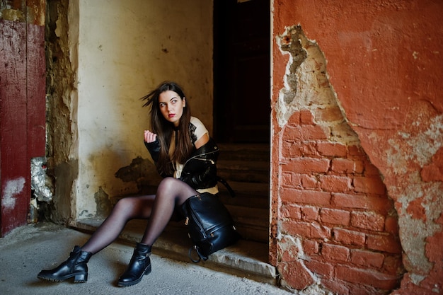 Stylish brunette girl wear on leather jacket and shorts with backpack sitting on old stairs against orange wall