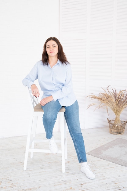 Стильная брюнетка плюс размер модельная женщина позирует, сидя на стуле в студии на белом фоне, портрет в полный рост.