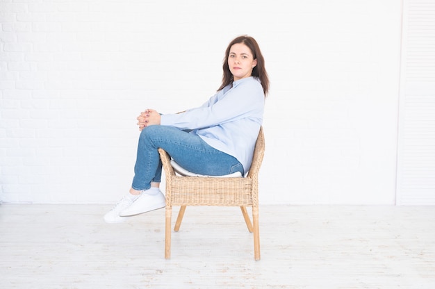 흰색 바탕에 스튜디오의 의자에 앉아 포즈를 취하는 세련된 브루네트 플러스 사이즈 모델 여성.